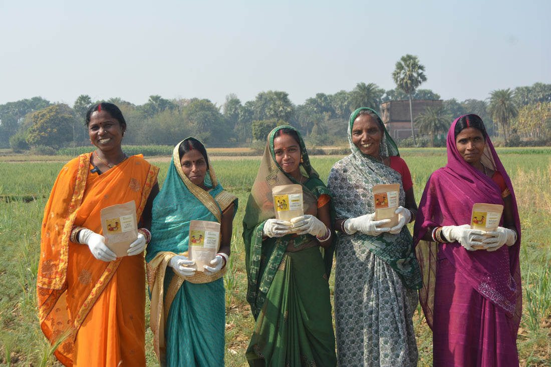 Five smiling Indian women farmers standing in a rural field wearing colorful saris and white safety gloves for clean, high-quality food processing, proudly holding packaged Natureship products.